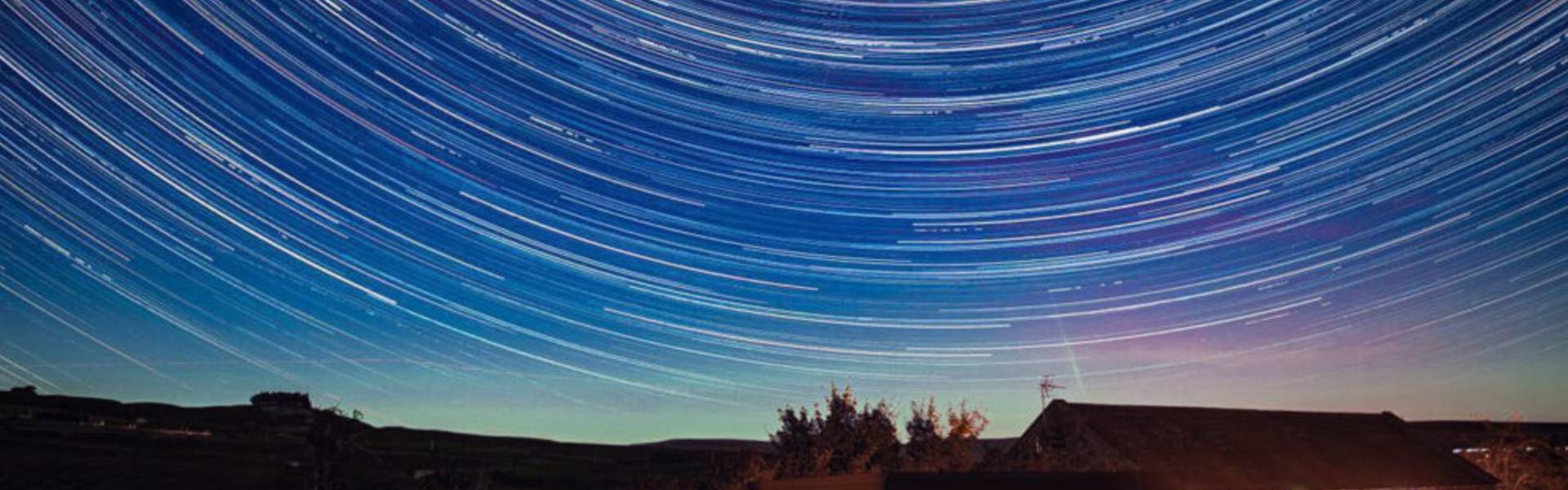 Long-exposure photograph showing circular star trails across a clear night sky above Grassholme Observatory, with the illuminated observatory building and surrounding countryside below.